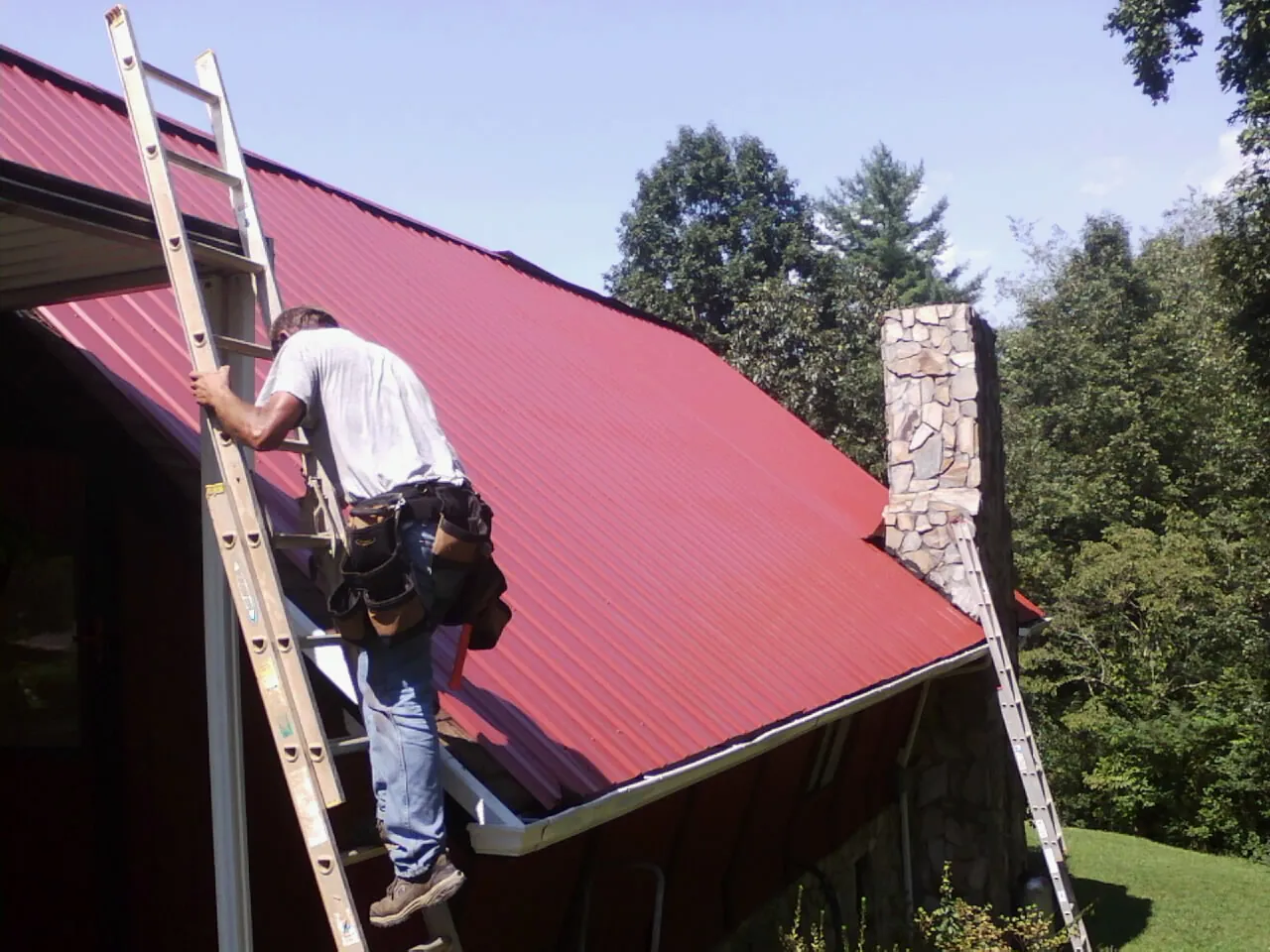 Roofer on a ladder beside a red metal roof and stone chimney — Soffit & Fascia Repair by Roofing Brothers in East Hanover